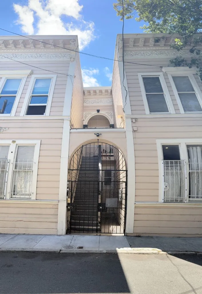 Beige wooden exterior, arched entrance in the center, black wrought iron gate. Stairs visible inside.