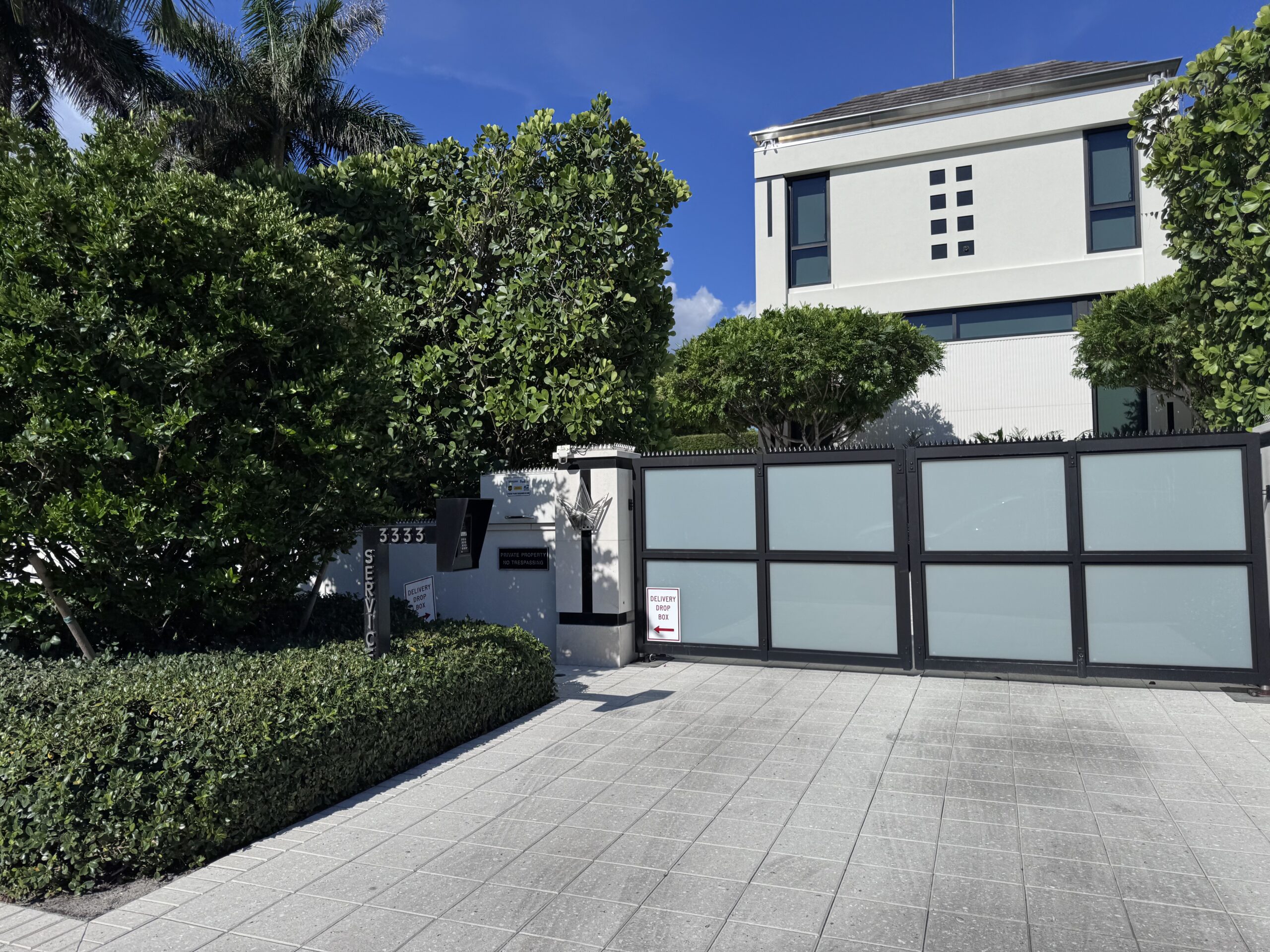 Maison blanche moderne derrière un portail coulissant en verre dépoli à cadre noir. Entourée d'arbres et de haies sous un ciel bleu.