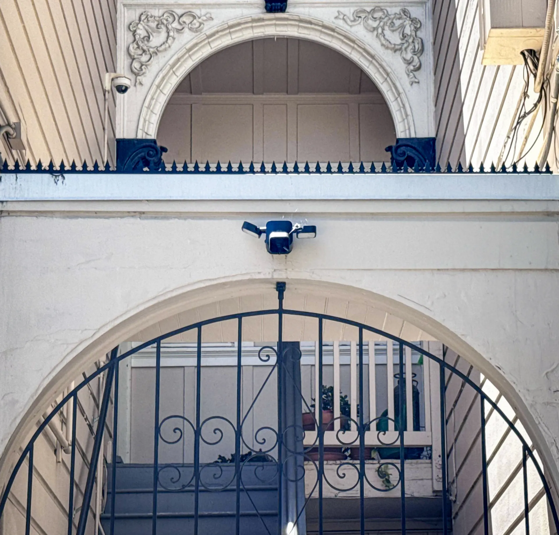 Decorative plaster arch with anti-climb spikes along the ledge, a security camera, and a scroll-patterned wrought iron gate below.