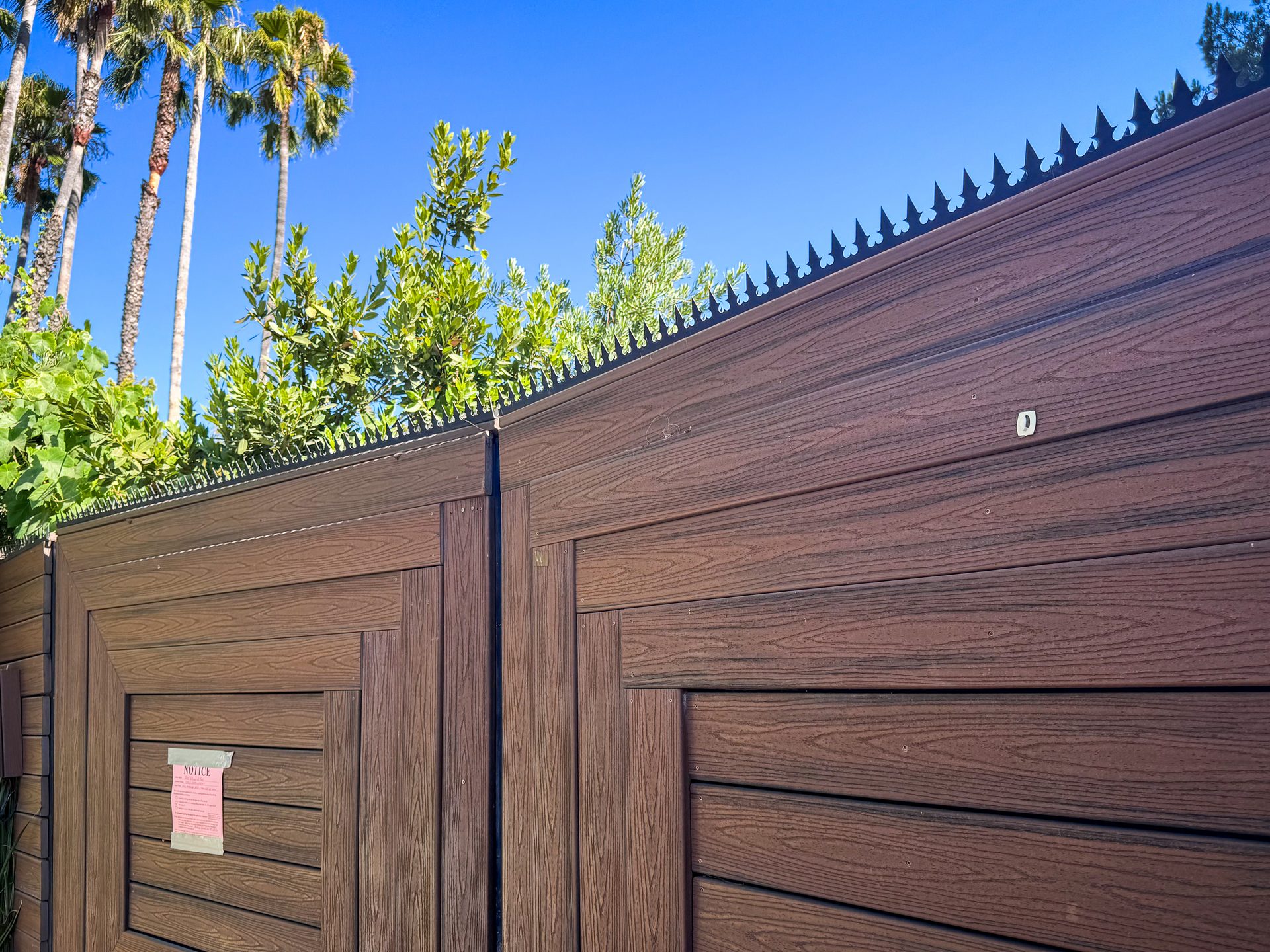 Vista geral dos espigoes anti-intrusao instalados de forma continua no topo de um portao/cerca alto com textura de madeira, com palmeiras ao fundo