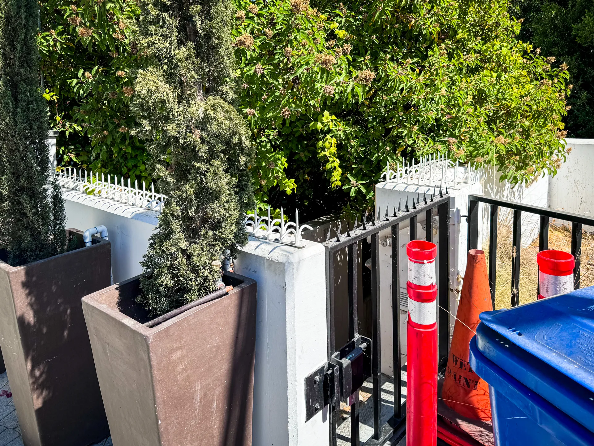 Spikes line the white wall around the entrance. A small black gate, planters, and safety cones.