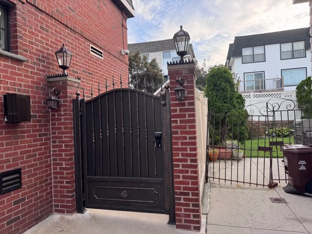 Black metal pedestrian gate between red brick pillars, residential carport with anti-climb spikes and lantern lighting above