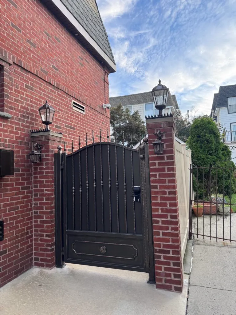 Angled view of black iron gate between brick pillars with lanterns, showing spiked security detail at the top edge