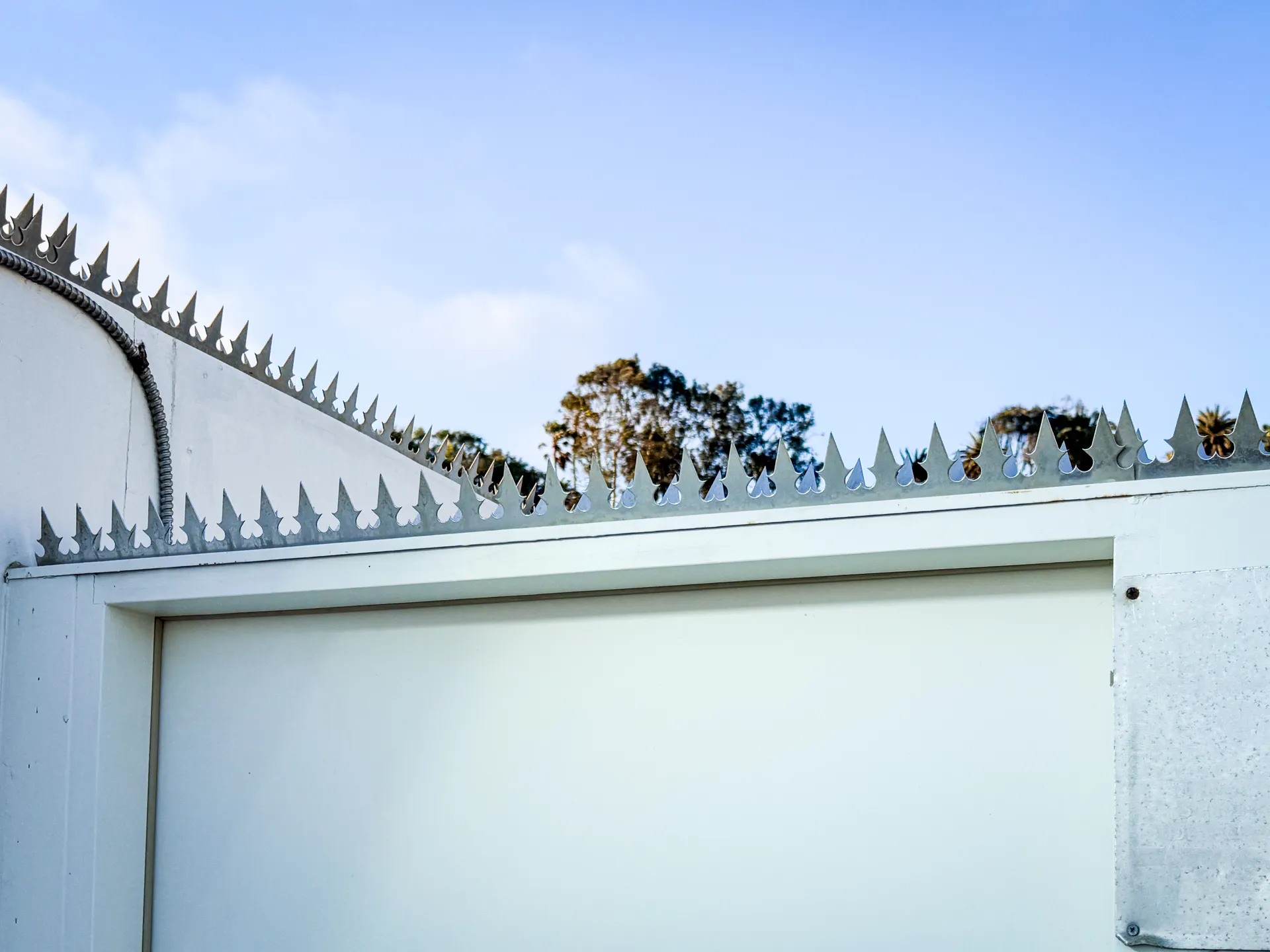 White sliding gate panel topped with anti-climb spikes, palm trees and warning signage against a clear sky
