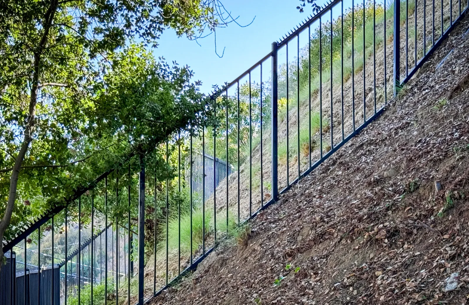 Black metal fence with top spikes running along a steep slope under trees.