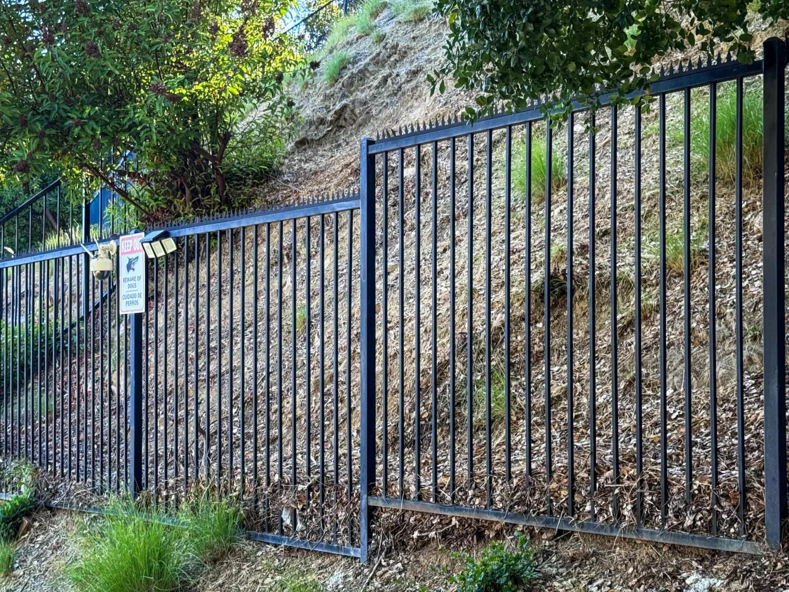 Black perimeter metal fence on sloping terrain. Spikes on the top edge, with trees and warning signs around.
