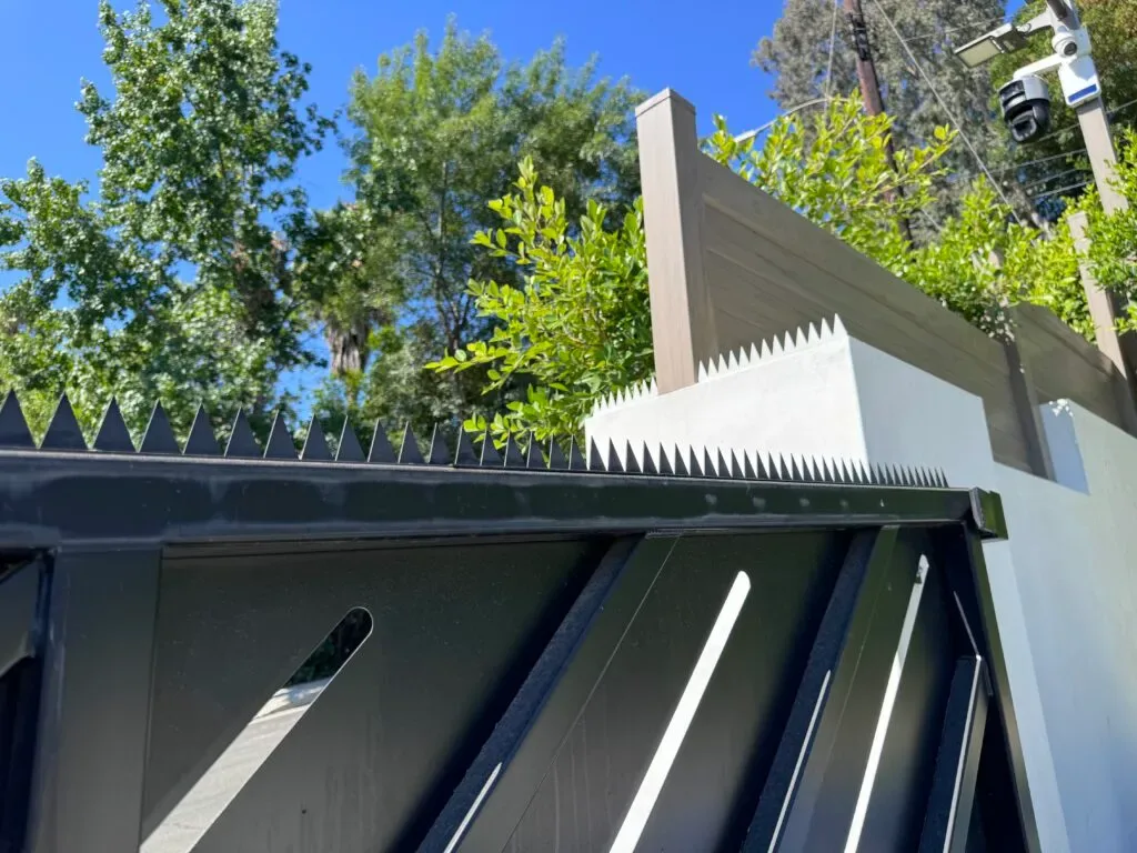Nahaufnahme eines schwarzen Tors mit dreieckigen Spikes neben einer weißen Wand und einem hölzernen Sichtschutzpaneel.