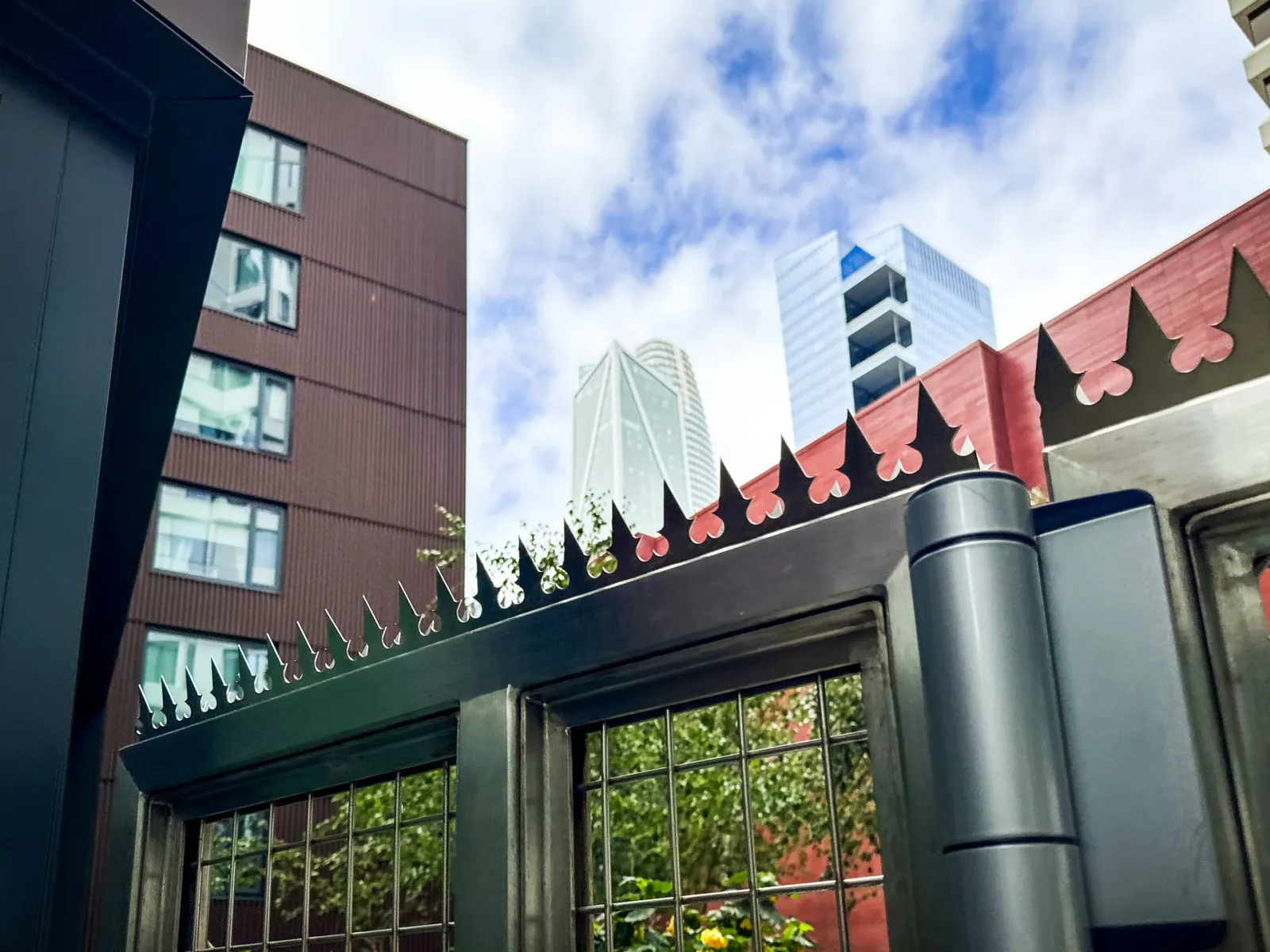 Silberfarbene Gothic-Anti-Kletter-Spikes mit Herzmotiv entlang der Oberkante eines dunklen Metalltors an der Rückseite, im Hintergrund Hochhäuser von San Francisco.