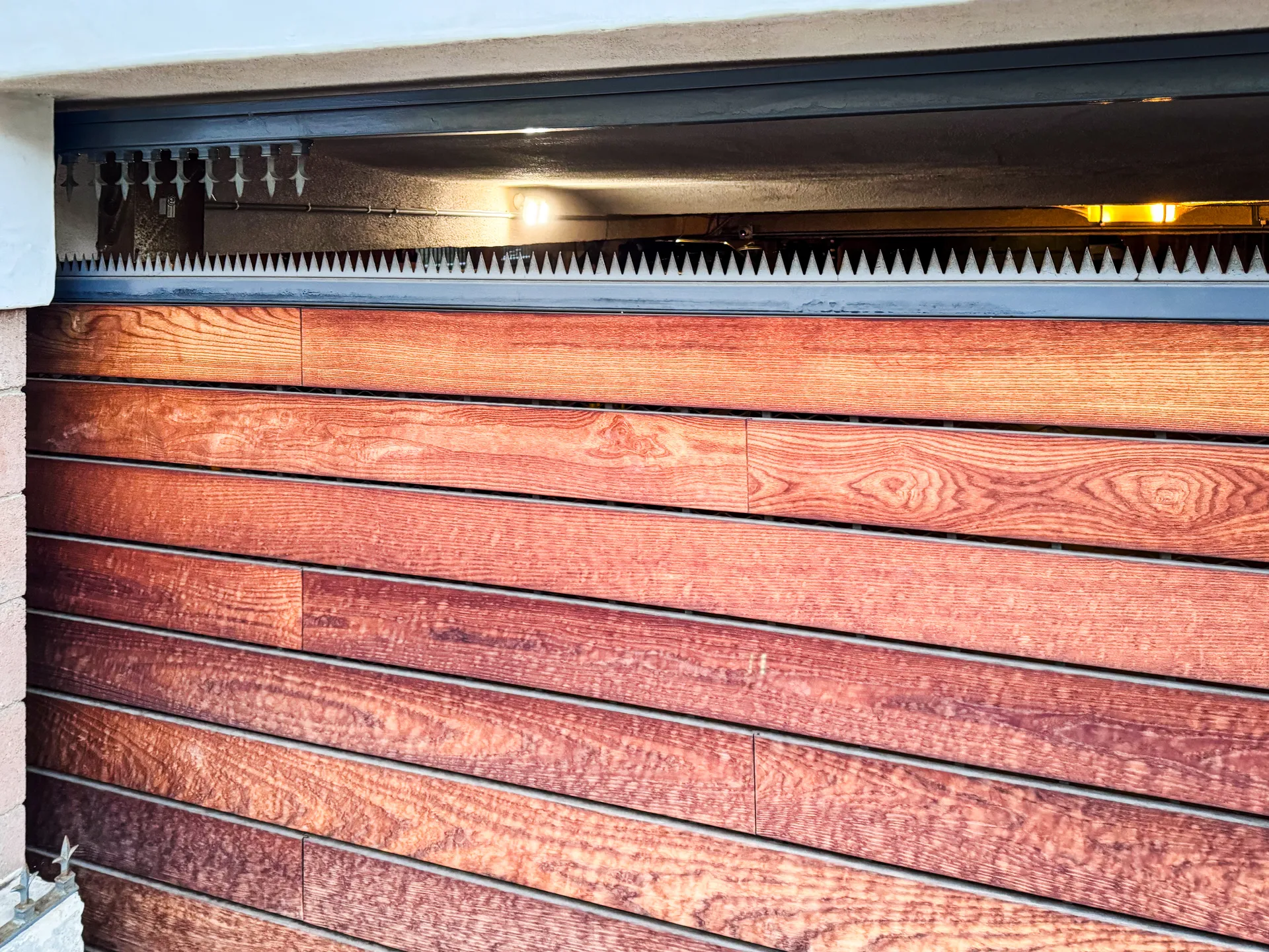 Mahogany horizontal-slat sliding driveway gate with anti-climb spikes along the top rail, set in a black steel frame