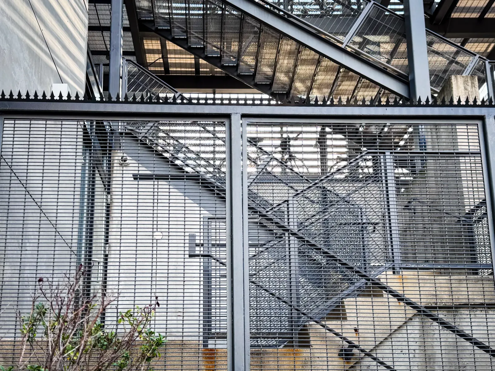 Tela de malha de aço a envolver uma escadaria, coroada com espigões anti-escalada pretos ao longo da aresta superior.
