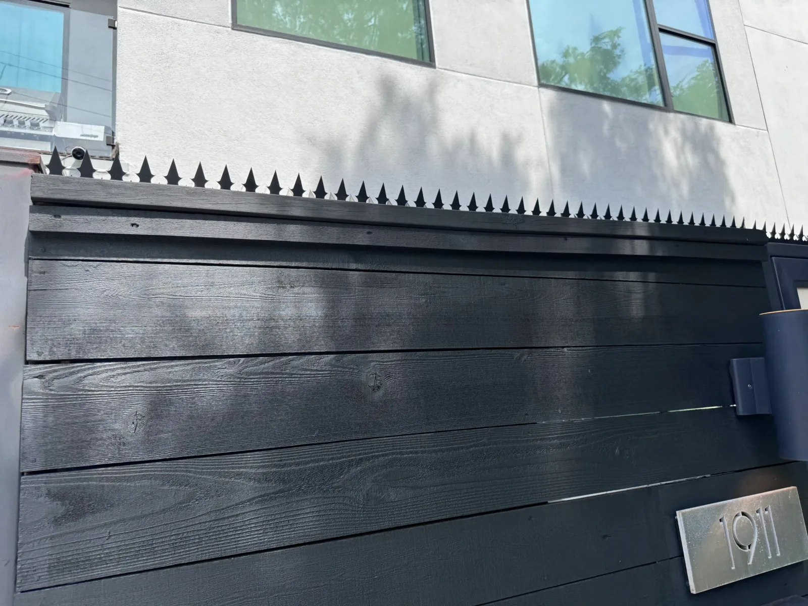 Hausnummernschild an einem Eckabschnitt mit schwarzem Zaun und Anti-Kletter-Spikes.