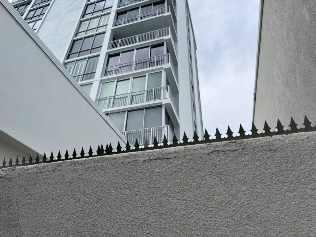 Close-up of black anti-climb spikes installed along a concrete and stucco boundary wall.