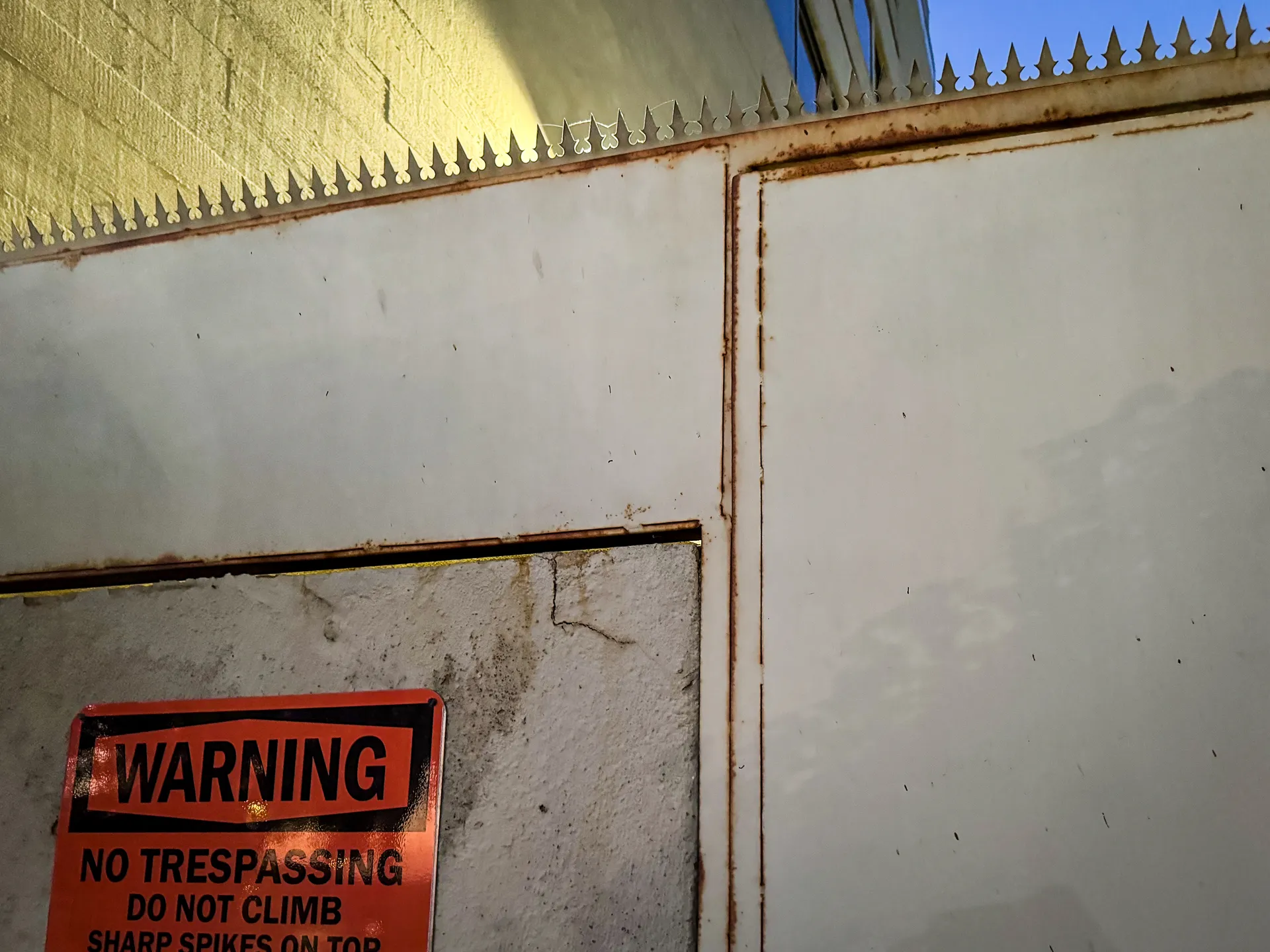 Painted concrete perimeter wall with anti-climb spikes along the top and a "No Trespassing — Sharp Spikes" warning sign, multi-story building behind
