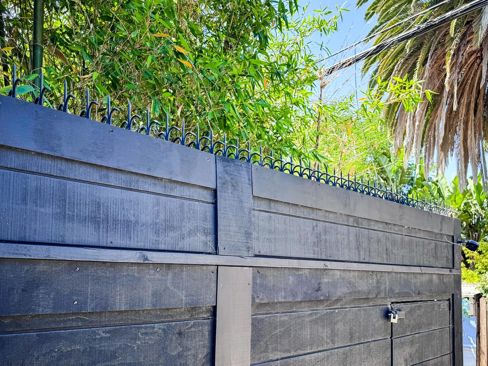 Black wood fence and gate corner with trees behind and a continuous spike line along the top.