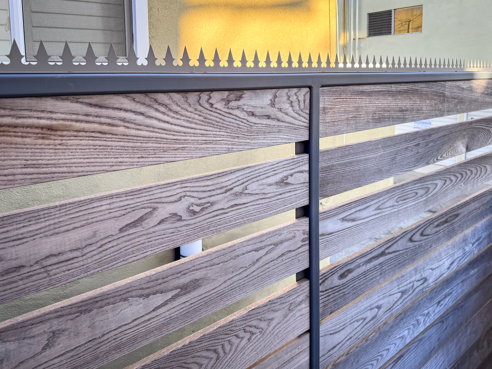 Gray wood-slat fence with brass accent strips and anti-climb spikes on a dark steel frame, stucco building and security camera above
