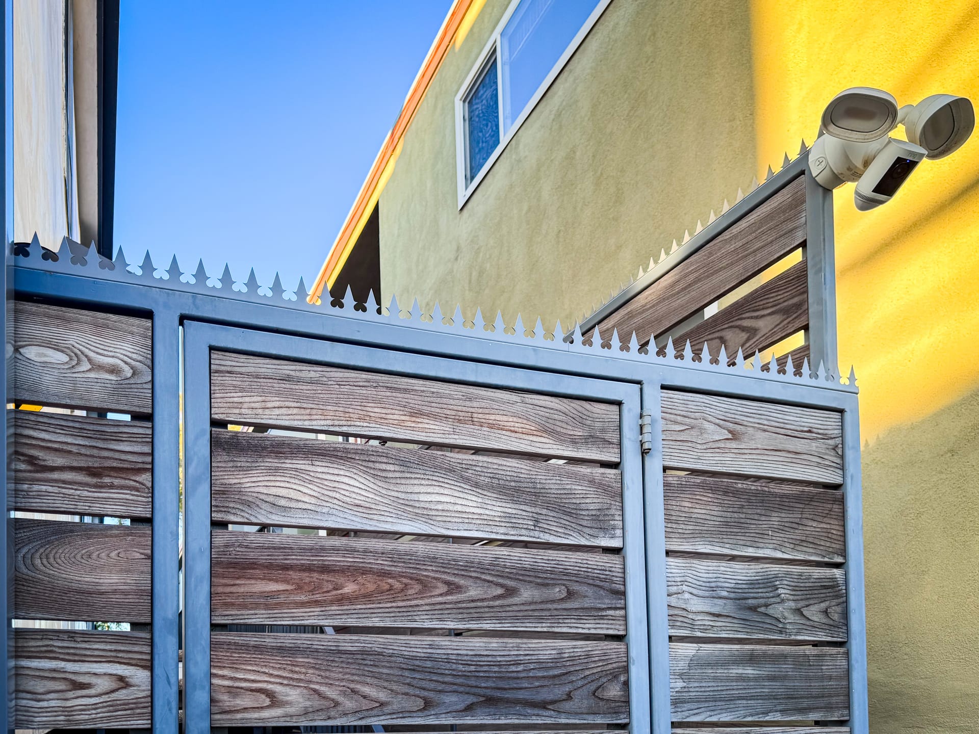Wood-Slat Privacy Fence & Gate — Anti-climb spikes integrated into a gray wood-slat privacy fence and pedestrian gate with custom brass accents, hardenin
