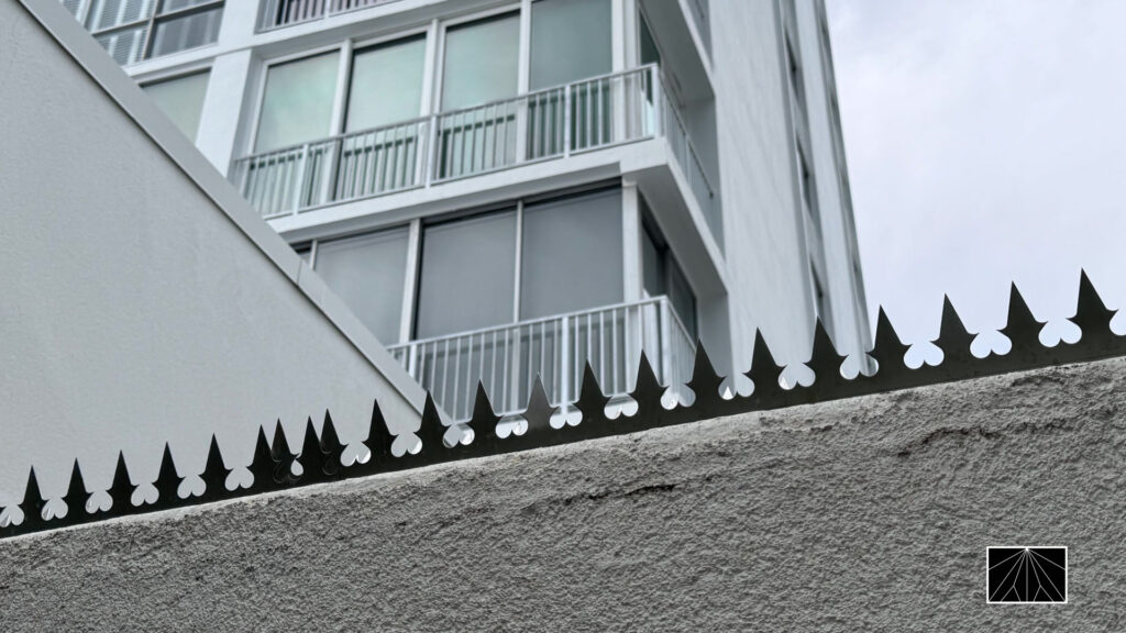 Black anti-climb security spikes installed on top of a concrete wall with a modern apartment building in the background