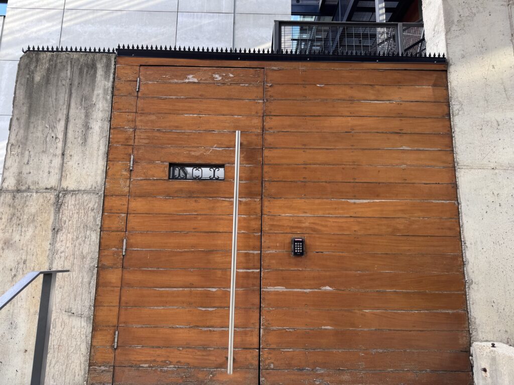 Tall wood entry gate between concrete wing walls, topped with a continuous black GIZAGIZA HEART crown.