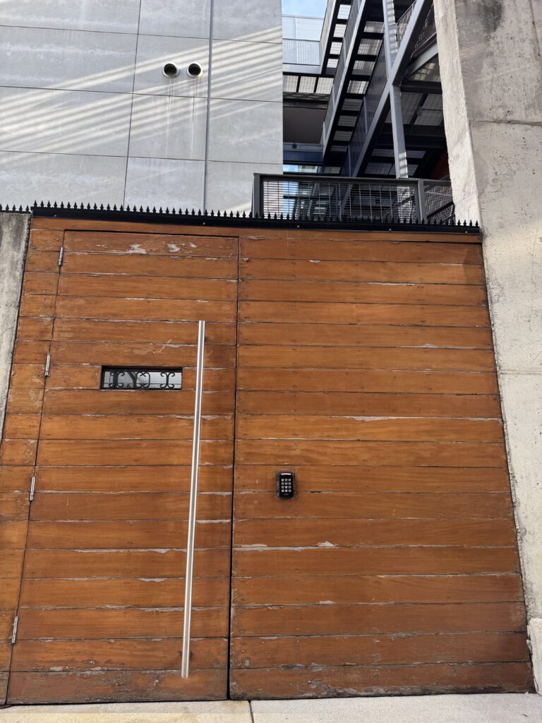 コンクリートの袖壁に挟まれた背の高い木製ゲート。上部をブラックの GIZAGIZA HEART が連続して冠する。