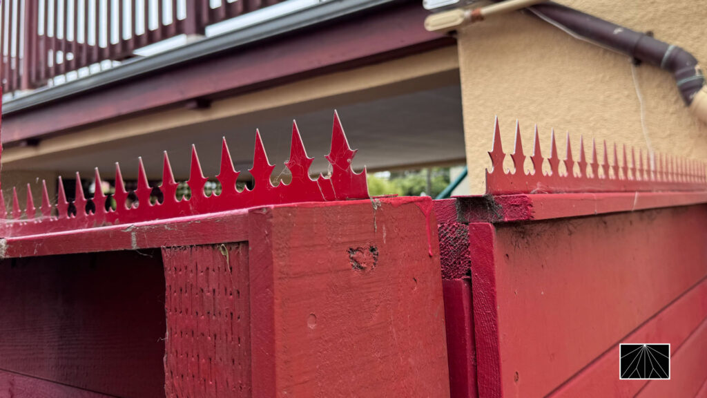 Red fence topped with gothic-style anti-climb spikes next to a stucco wall and exterior pipes.