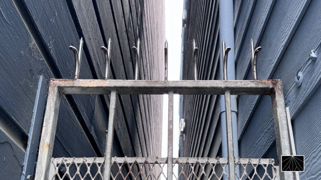 Close-up of anti-climb spikes on a steel mesh gate between dark exterior walls