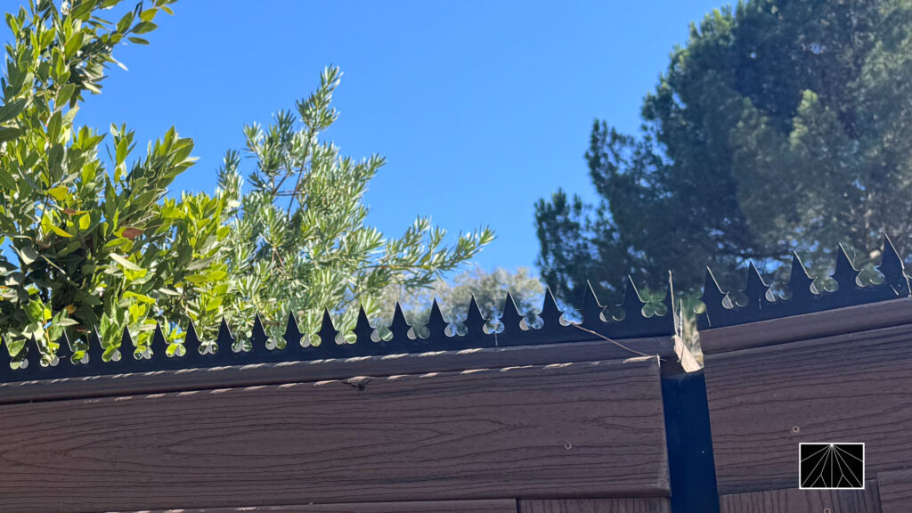 Close-up of black anti-climb security spikes mounted on the top of a wood-look fence against blue sky and greenery