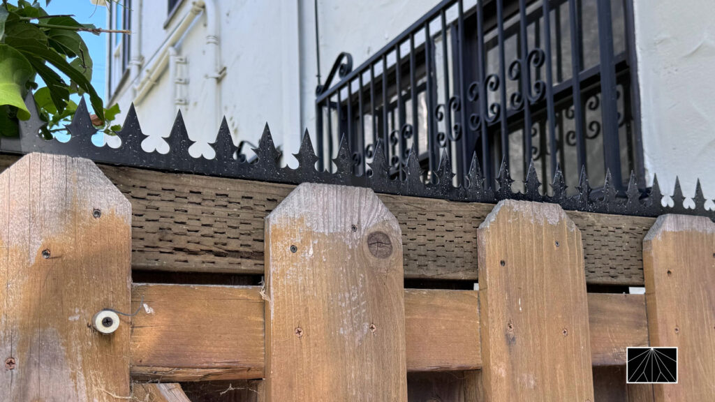 Close-up of decorative anti-climb security spikes mounted along the top of a wooden fence, with a window security grille in the background.