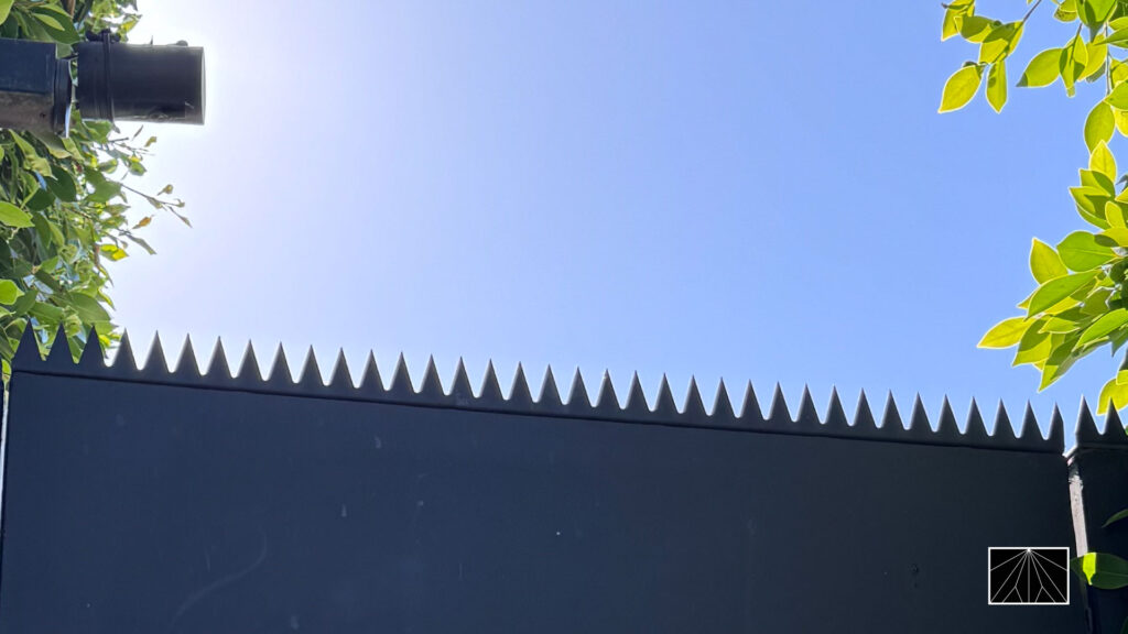 Close-up of anti-climb security spikes installed along the top edge of a black metal gate, framed by hedge leaves against a clear blue sky.