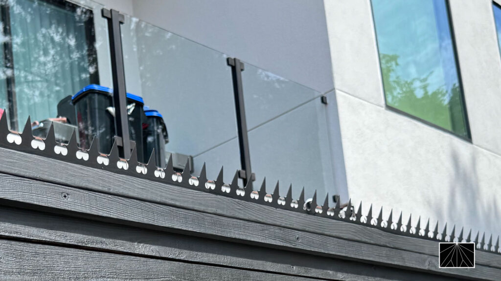 Decorative anti-climb fence spikes on a black wooden fence below a glass balcony railing on a modern home