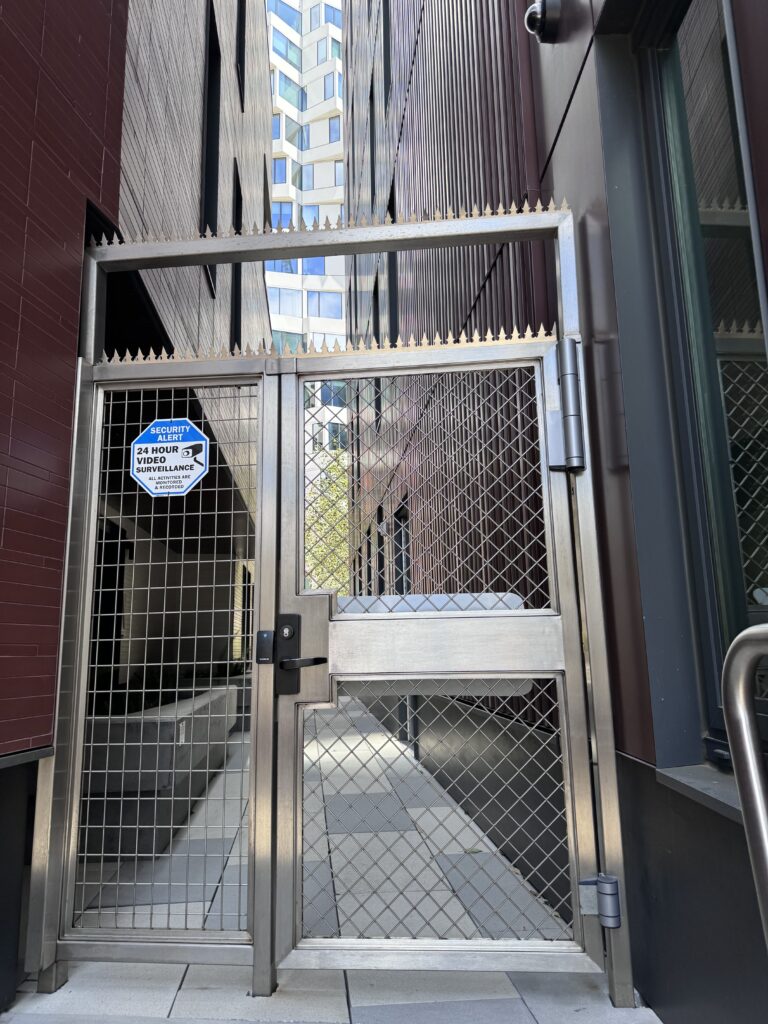 Ein modernes Sicherheitstor aus Metall mit scharfen Zacken an der Oberseite, das einen schmalen Durchgang zwischen modernen Gebäuden in San Francisco absperrt.