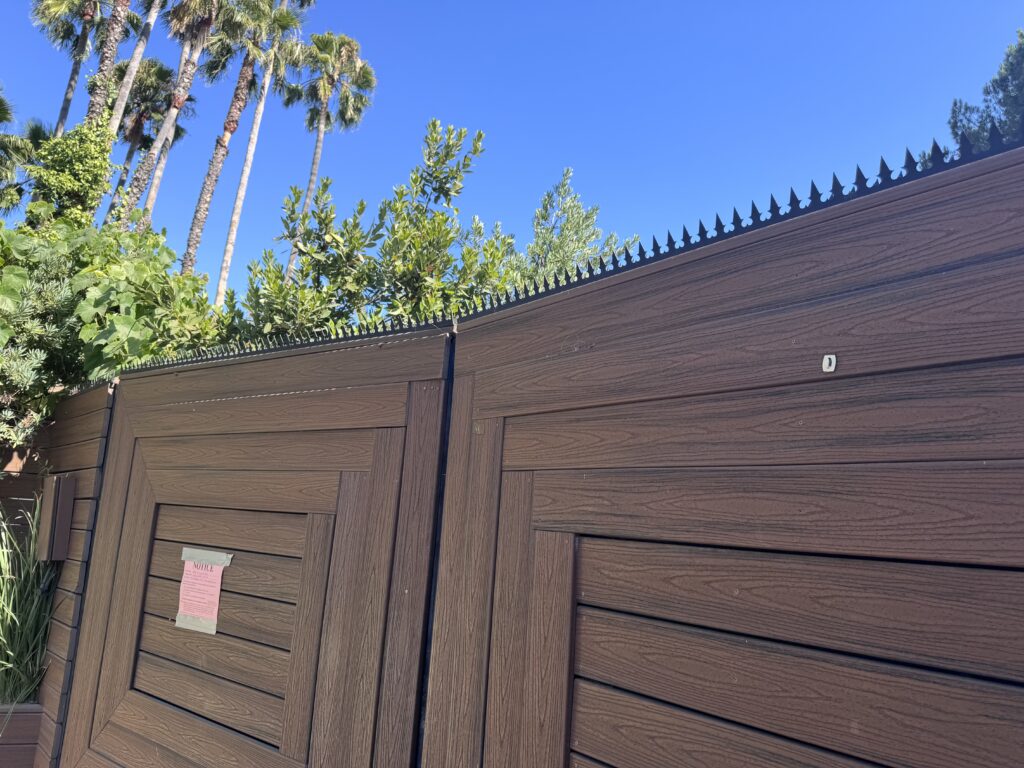 Wide view of a tall wood-look gate/fence topped with anti-climb security spikes, with palm trees in the background