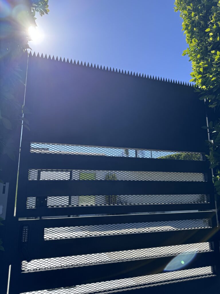 Upward view of anti-climb spikes along the top of a black metal gate, with sunlight flare.
