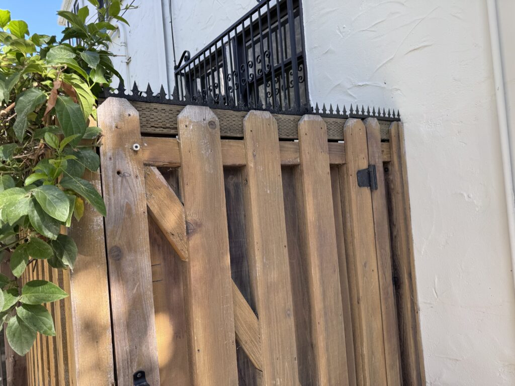 Wooden side gate with a reinforced frame, topped with anti-climb security spikes beneath a window grille.