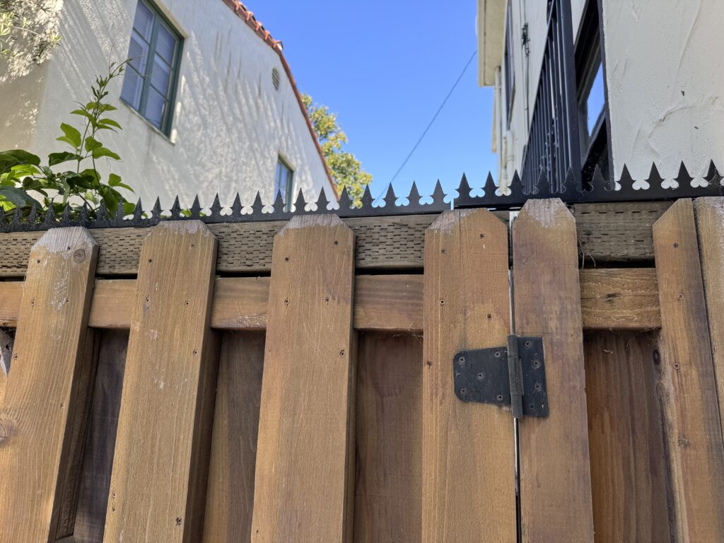 Fence-line close-up showing anti-climb security spikes spanning the top of the wooden gate and adjacent panels.
