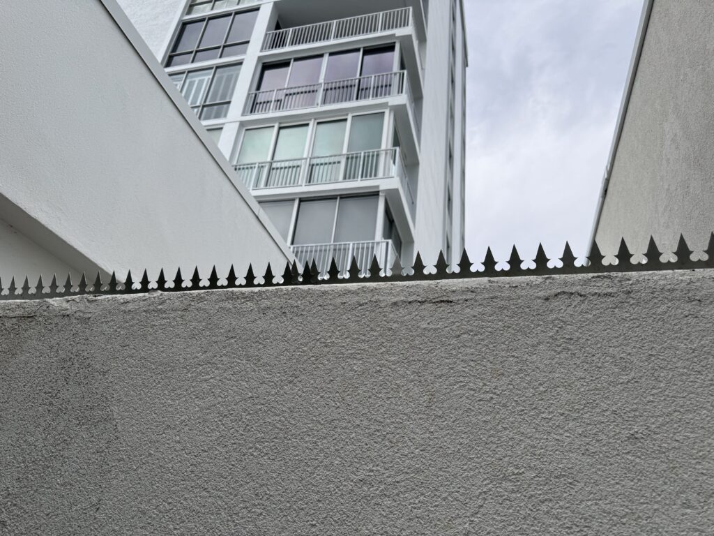 Púas de seguridad que recubren la parte superior de un muro fronterizo, vistas hacia arriba, en dirección a una torre de gran altura.