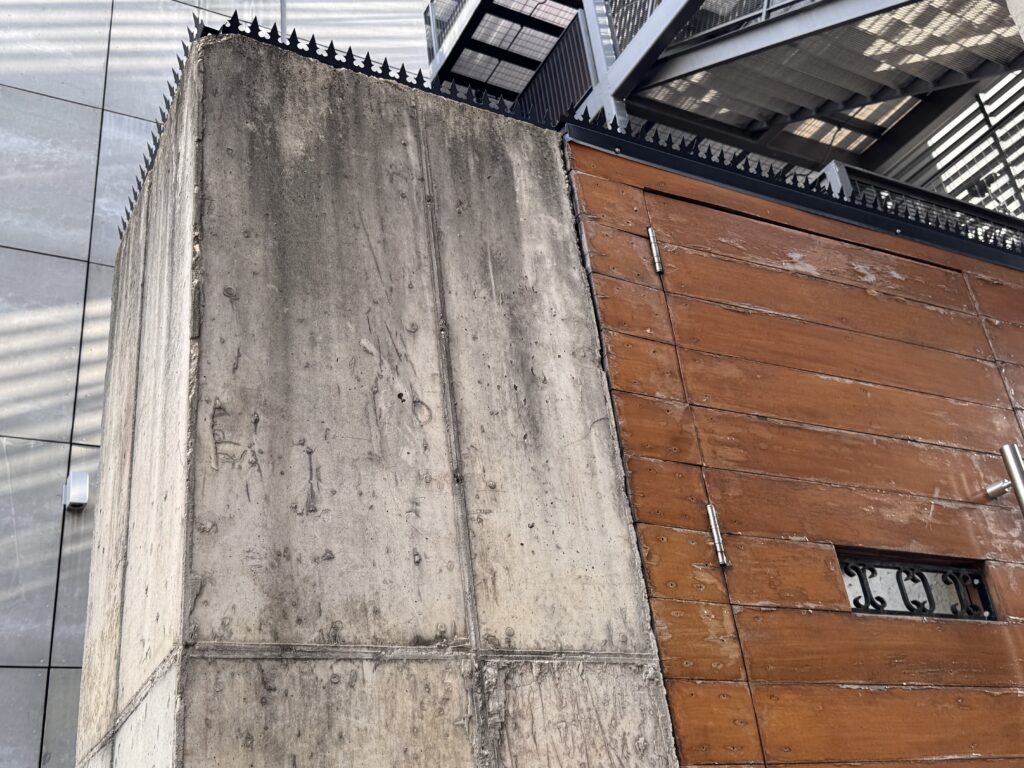 Unión entre fachada de hormigón bruto y puerta de madera con pequeña ventana decorativa.