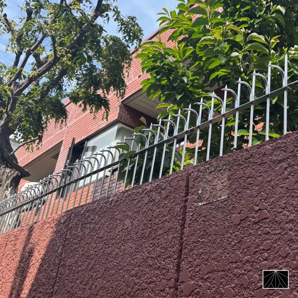 Barrera de seguridad Ninja Deterrent Classic con pinchos plateados curvados, instalada sobre un muro rojo texturizado bajo la luz del día, destacando su fuerte efecto disuasorio.