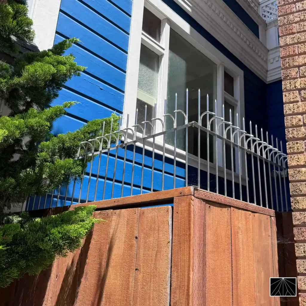 Barrera de seguridad Ninja Deterrent con pinchos plateados curvados instalada en la ventana y balcón de una casa azul, reforzando la protección contra intrusos.