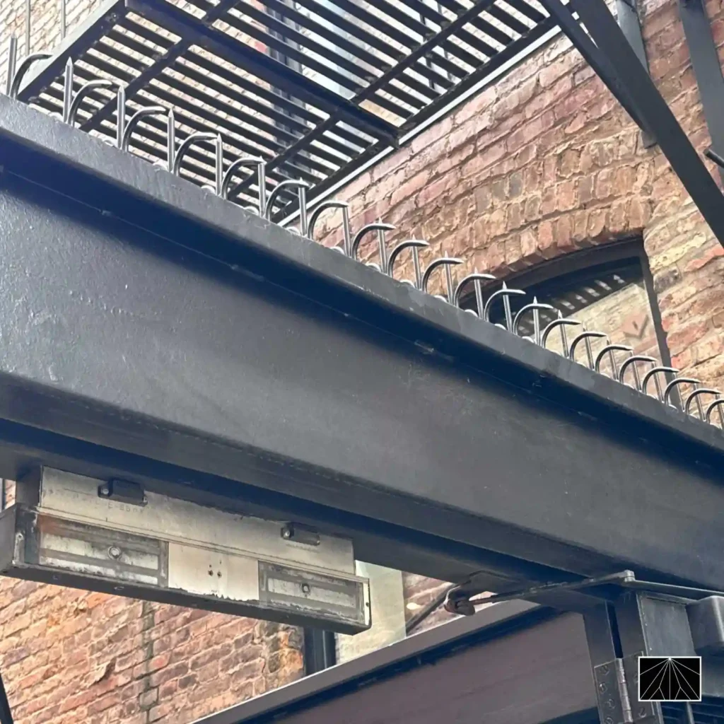 Ninja Deterrent Abwehrspitzen Classic in Schwarz an einem Stahlträger einer Feuertreppe zur Verhinderung von unbefugtem Aufstieg.
