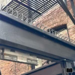 Ninja Deterrent Abwehrspitzen Classic in Schwarz an einem Stahlträger einer Feuertreppe zur Verhinderung von unbefugtem Aufstieg.