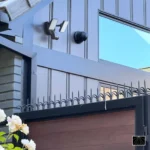 Barrera de seguridad Ninja Deterrent con pinchos decorativos instalada en un portón de garaje junto a un reflector de luz para maximizar la disuasión nocturna.