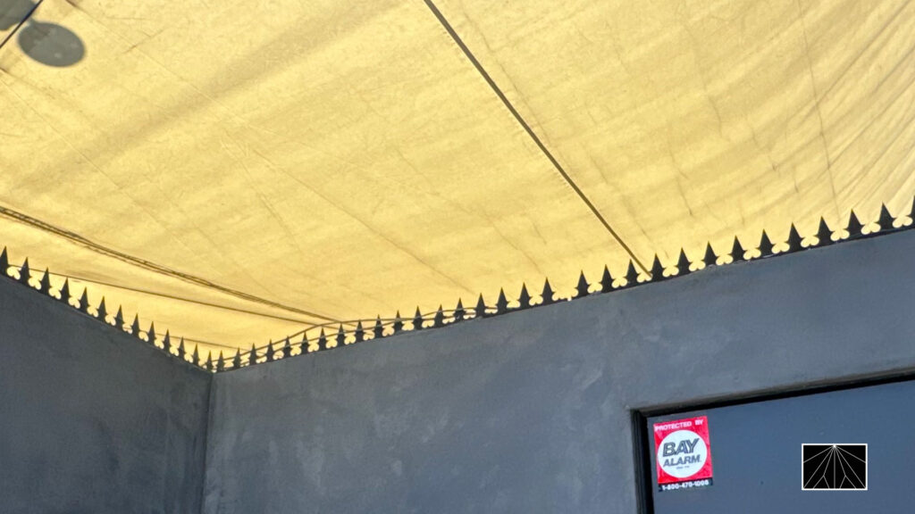Gothic-style anti-climb spikes running along the top edge of a gray courtyard wall beneath a canopy, beside a doorway with a security alarm sticker.