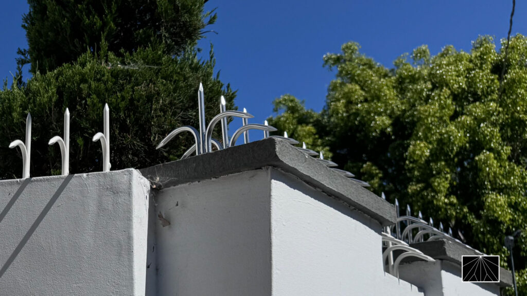 White perimeter wall with silver curved anti-climb spikes along the top edge, framed by trees and blue sky.