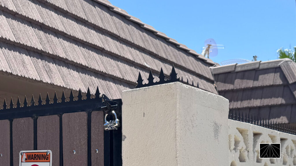 Gothic anti-climb spikes installed along the top of a black gate and a white stucco pillar, with a warning sign visible below under a clear blue sky.
