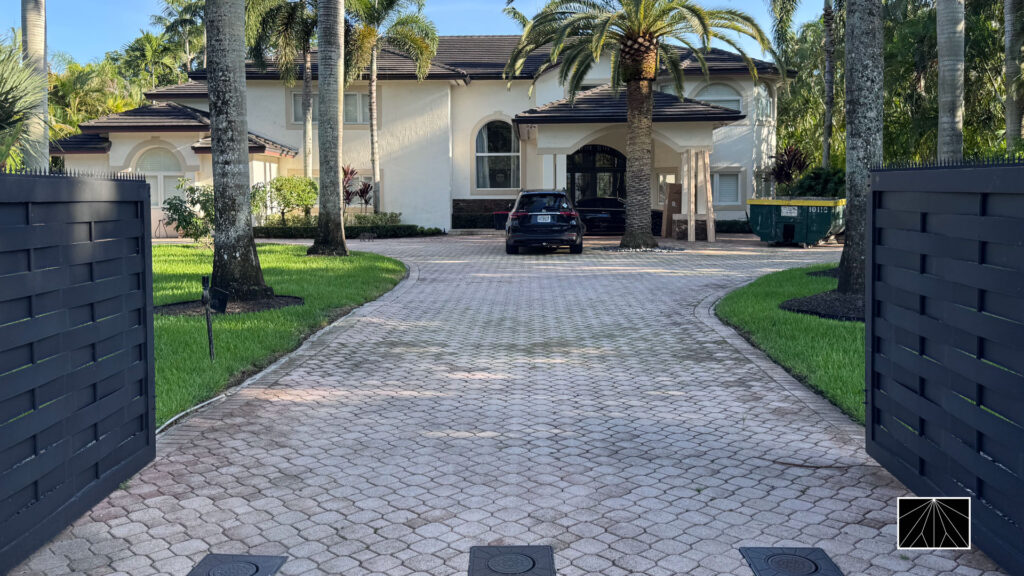 Open black driveway gates with spear-top anti-climb spikes framing a palm-lined paver drive leading to a large white house.