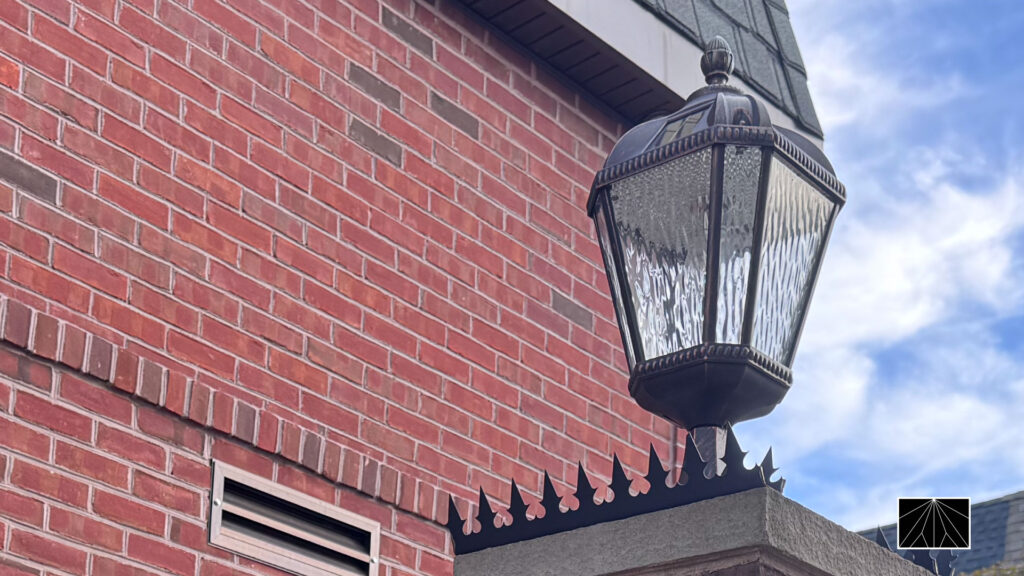 Gothic anti-climb spikes mounted on a stone-capped brick pillar beneath a lantern light on a red brick home.