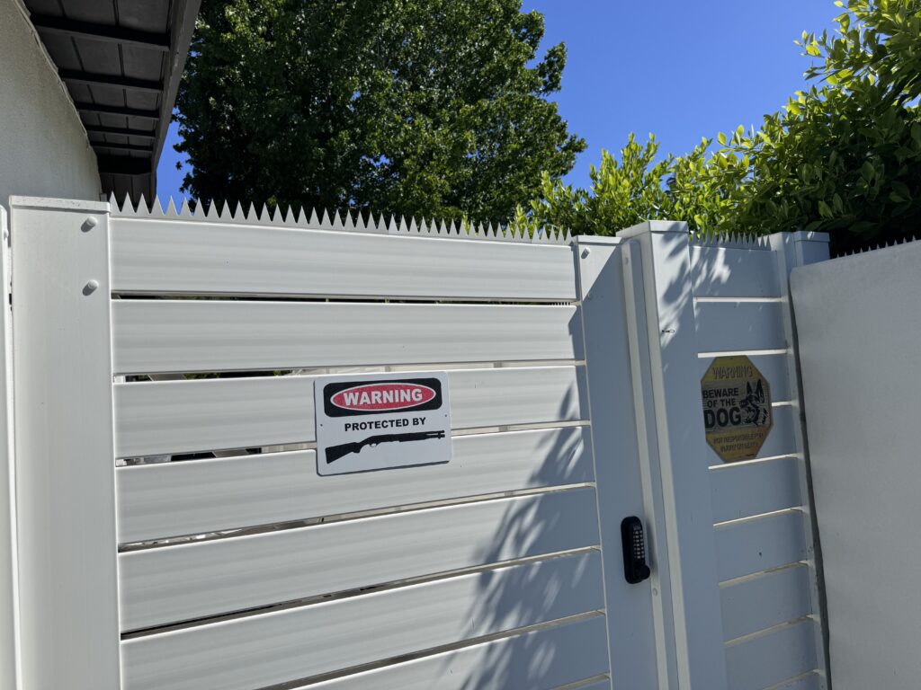 Portão lateral branco com espigões antiescalada no topo, com placa de aviso fixada no painel