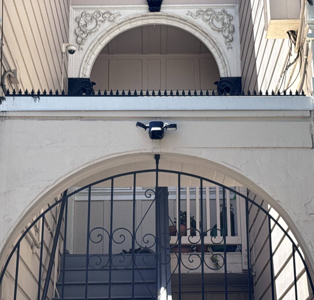 Primer plano del arco de entrada con púas anti-escalada y una cámara de seguridad; detrás de la reja se ve la escalera.