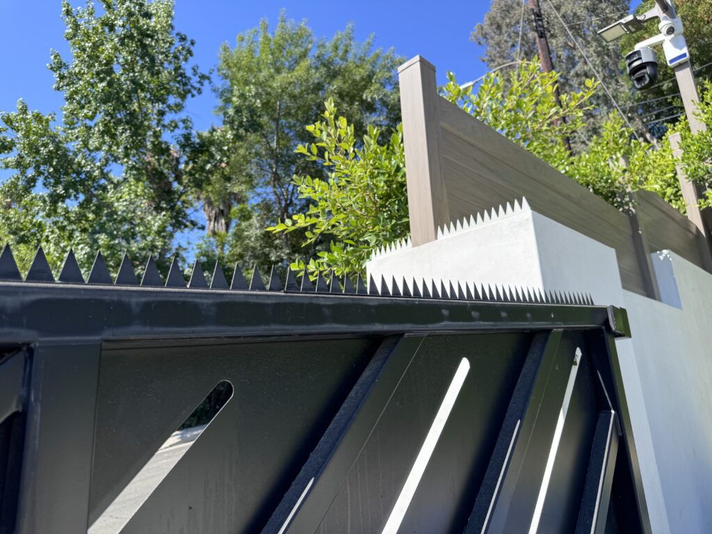 Close-up eines schwarzen Tores mit dreieckigen Anti-Climb-Spikes, neben einer weißen Wand und Holz-Sichtschutzelementen