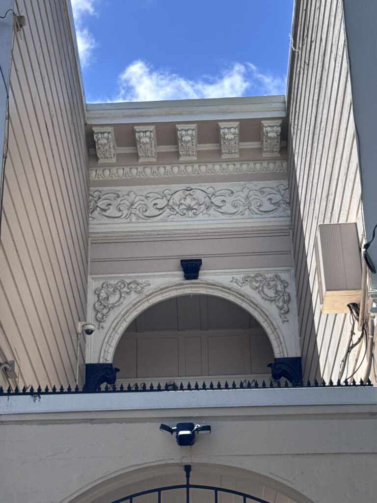 Vista para cima de um arco e cornija ornamentados, emoldurados por paredes laterais; pontas anti-escalada visíveis sob o céu azul.